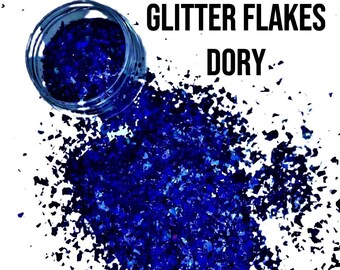 Op de afbeelding: Blauwe glittervlokken in een pot, die uitstrooien op een witte achtergrond. De pot heeft een doorzichtig deksel en de tekst "GLITTER FLAKES DORY" is op de witte achtergrond gedrukt.