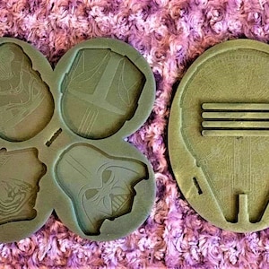 Könnte beinhalten: Eine grüne Silikonform mit fünf verschiedenen Formen. Die Formen sind von Star Wars-Charakteren, darunter Darth Vader, Kylo Ren und ein Stormtrooper. Die Form befindet sich auf einer rosa und weißen flauschigen Oberfläche.