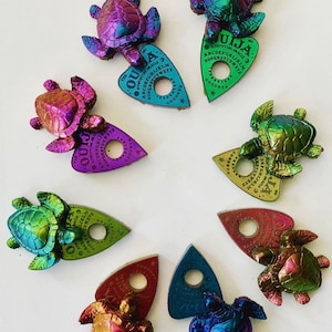 May include: Seven iridescent green, blue, purple, and red turtle charms with a Ouija planchette attached to each. The planchette is a triangular shape with a hole in the center and the word "Ouija" printed on it.