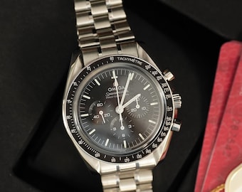 May include: An Omega Speedmaster Professional watch with a black dial, three sub-dials, and a stainless steel link bracelet. The bezel features a tachymeter scale. The watch face displays the Omega logo and Speedmaster Professional text.