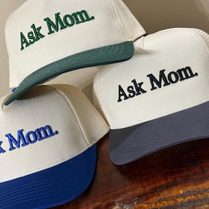 May include: Three cream-colored trucker hats with mesh backs. Each hat features the embroidered text "Ask Mom." in a different color: one with green text and a green brim, one with black text and a dark gray brim, and one with blue text and a blue brim.