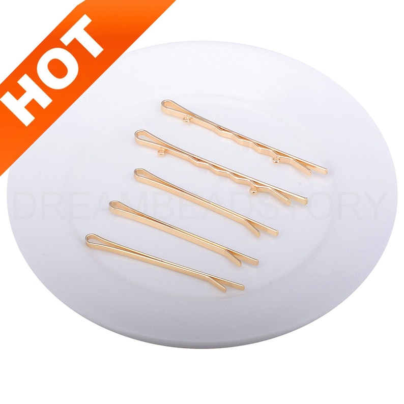 May include: Six gold metal hair clips on a white plate. The clips are different sizes and have a wavy design.