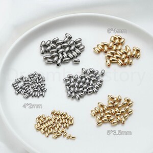 May include: An assortment of metallic oval beads in silver and gold tones, displayed on a white surface. The beads vary in size, with labels indicating dimensions such as 6*4mm, 5*3.5mm, and 4*2mm. These beads are suitable for jewelry making and crafting.