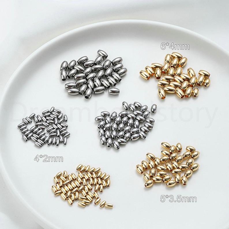 May include: An assortment of metallic oval beads in silver and gold tones, displayed on a white surface. The beads vary in size, with labels indicating dimensions such as 6*4mm, 5*3.5mm, and 4*2mm. These beads are suitable for jewelry making and crafting.
