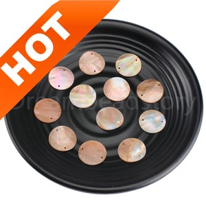 May include: A collection of 12 round, iridescent, mother-of-pearl shell beads with two holes each. The beads are arranged on a black plate.