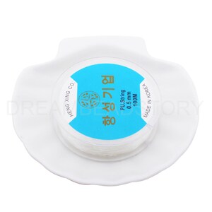Puede incluir: Una bobina blanca de cuerda de PU de 0,5 mm, etiquetada "항성기업" en coreano, con un fondo azul y el texto "PU String 0.5 mm 100M" en ingl&eacute;s. La bobina est&aacute; en un plato de cer&aacute;mica blanco.