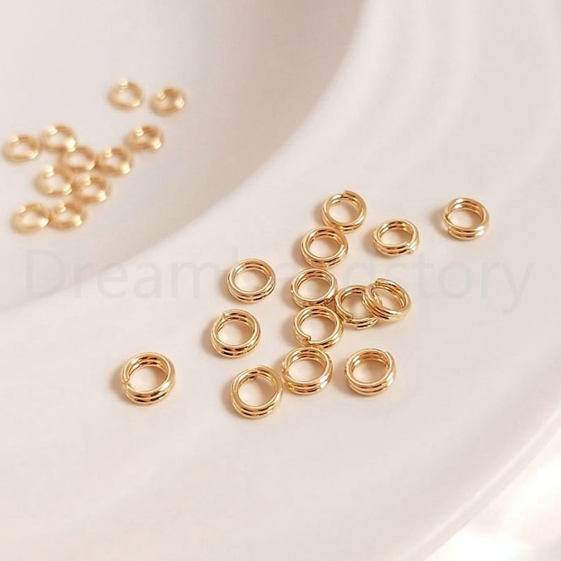 以下が含まれることがあります： 金色のジャンプリングのグループのクローズアップ。リングは小さく、滑らかで磨かれた仕上げになっています。ジュエリー作りやその他のクラフトプロジェクトに最適です。