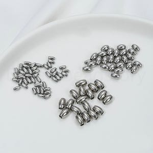 May include: A collection of shiny, silver-colored metal beads in various sizes and quantities. The beads are oval-shaped and appear to be made of stainless steel. They are arranged on a white surface, likely for jewelry making or crafting.
