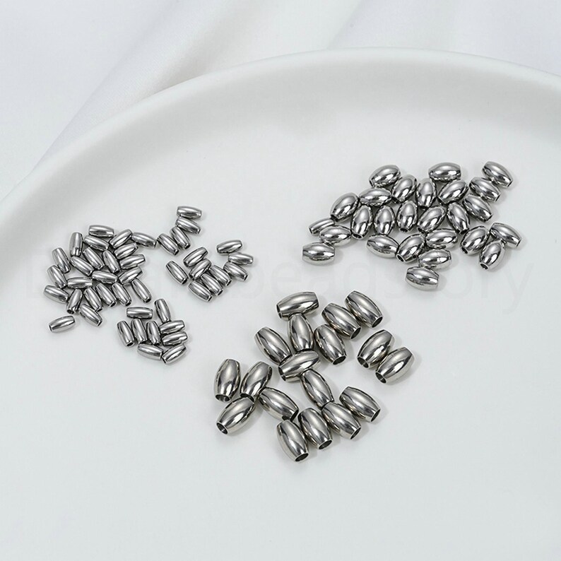 May include: A collection of shiny, silver-colored metal beads in various sizes and quantities. The beads are oval-shaped and appear to be made of stainless steel. They are arranged on a white surface, likely for jewelry making or crafting.