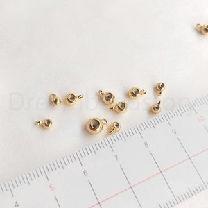 Puede incluir: Una colección de pequeños dijes de joyería dorados, con un cuerpo redondo y un pequeño lazo para sujetar. Los dijes tienen un área pequeña y empotrada en el centro. Una regla está en el fondo.