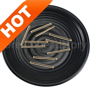 Könnte beinhalten: Nahaufnahme einer schwarzen Platte mit zehn goldenen Metall-Stangenverbindern. Die Verbinder sind etwa 2,5 cm lang und haben eine glatte, glänzende Oberfläche.