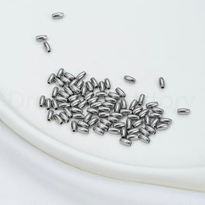 May include: A collection of small, shiny, silver-colored metal beads. The beads are oval-shaped and scattered on a white surface. These beads are likely used for jewelry making or other craft projects.