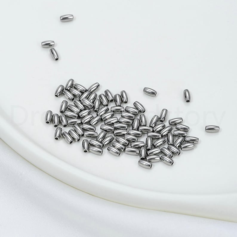May include: A collection of small, shiny, silver-colored metal beads. The beads are oval-shaped and scattered on a white surface. These beads are likely used for jewelry making or other craft projects.