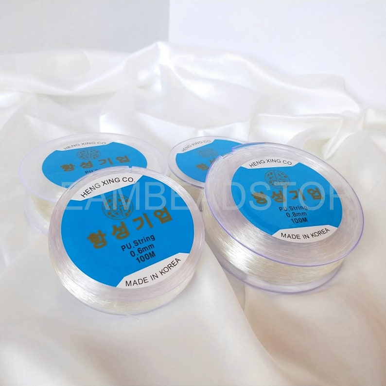 Puede incluir: Cuatro carretes de hilo el&aacute;stico transparente, cada uno con una etiqueta azul que dice "Heng Xing Co. 항성기업 PU String 0.6mm 100M Made in Korea" o "Heng Xing Co. 항성기업 PU String 0.8mm 100M Made in Korea".