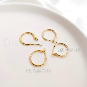 May include: Two pairs of gold hoop earrings on a white surface. The earrings are different sizes. The larger pair is labeled "14K Gold Color" and the smaller pair is labeled "18K Gold Color".