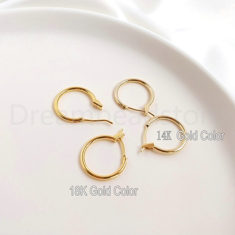 May include: Two pairs of gold hoop earrings on a white surface. The earrings are different sizes. The larger pair is labeled "14K Gold Color" and the smaller pair is labeled "18K Gold Color".