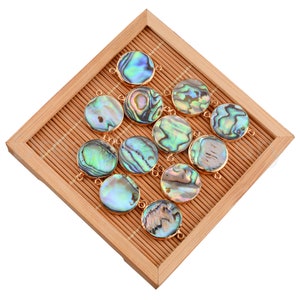 May include: A collection of 12 round abalone shell pendants with gold-toned metal accents. The pendants are arranged on a bamboo mat.