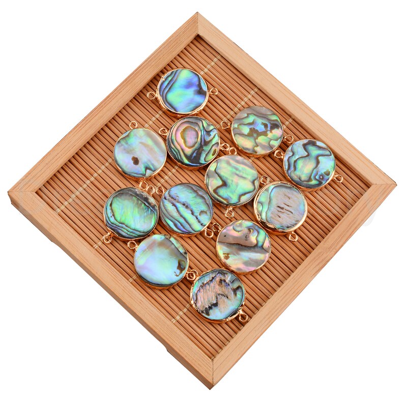May include: A collection of 12 round abalone shell pendants with gold-toned metal accents. The pendants are arranged on a bamboo mat.