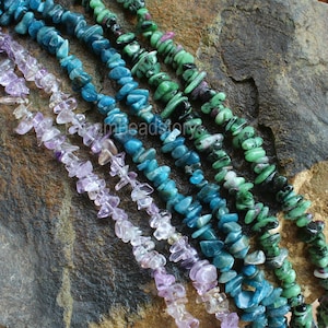 May include: Five strands of chip gemstone beads in various colors, including green, blue, and purple. The beads are arranged in rows on a dark gray textured surface.