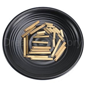 May include: A collection of small, rectangular brass bars with a single hole at one end, displayed on a black, textured plate. The bars vary slightly in size and are arranged in a scattered pattern.