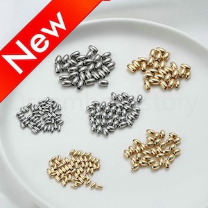 May include: An assortment of metallic oval beads in silver and gold tones. The beads are scattered on a white surface. A red banner in the upper left corner displays the word "New" in white.