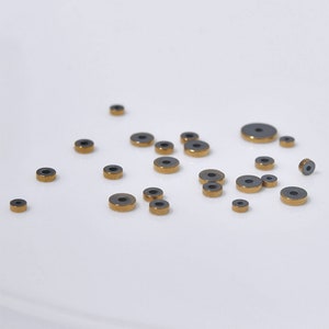 Pu&ograve; includere: Una collezione di piccoli anelli rotondi in ceramica nera con bordo dorato. Gli anelli sono sparsi su una superficie bianca.