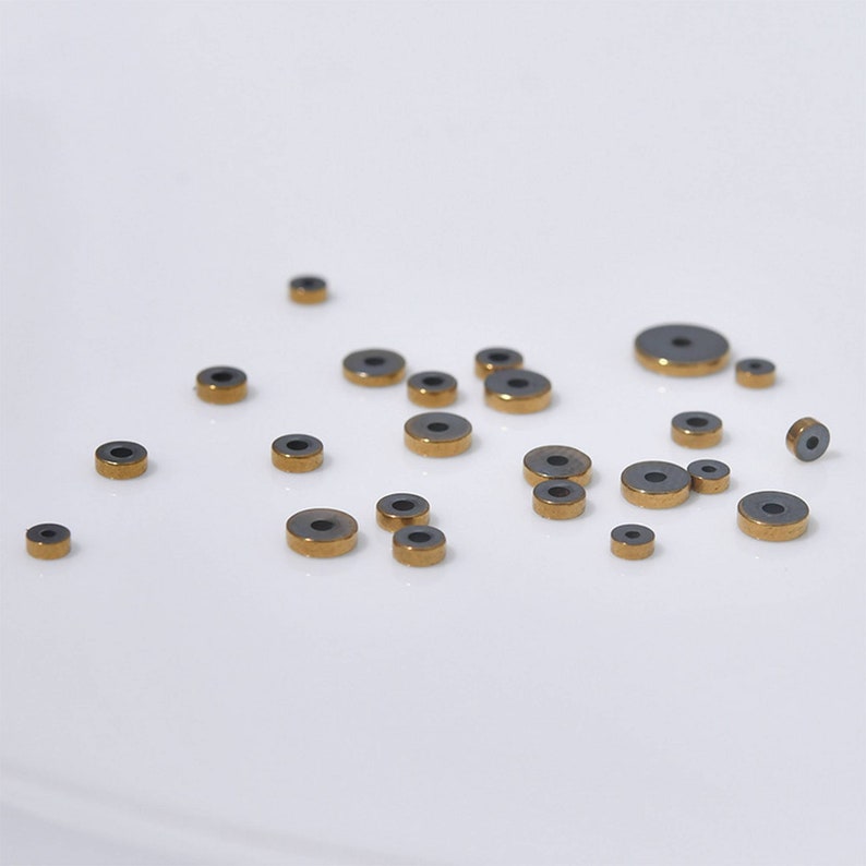 Pu&ograve; includere: Una collezione di piccoli anelli rotondi in ceramica nera con bordo dorato. Gli anelli sono sparsi su una superficie bianca.