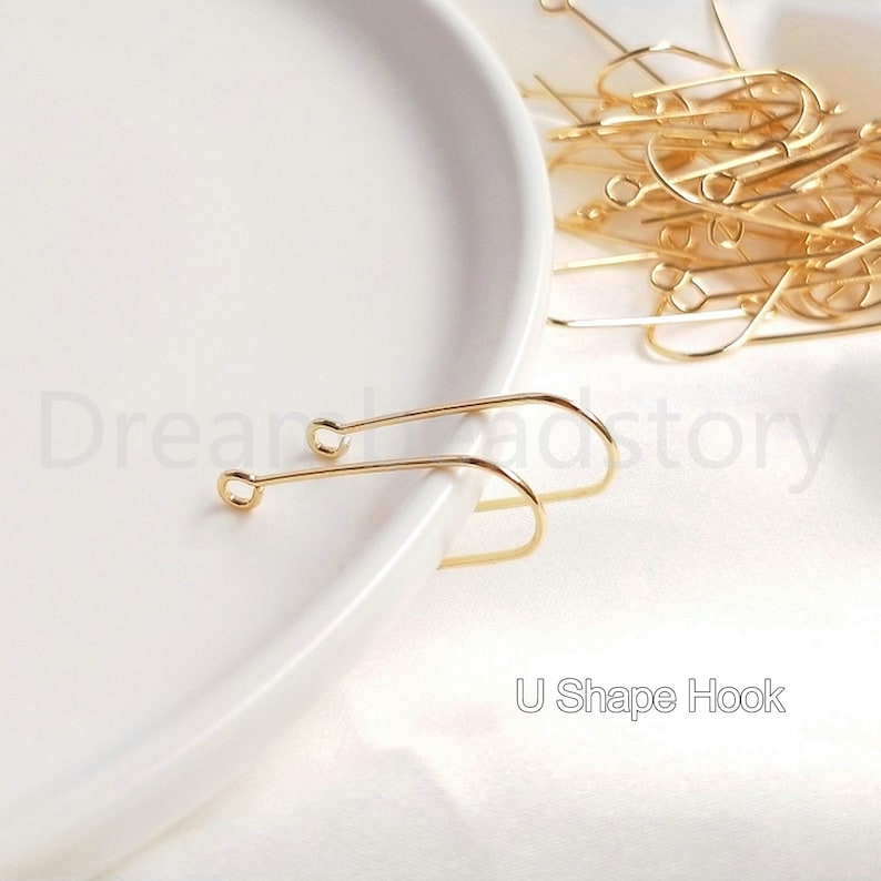 May include: Two gold U-shaped earring hooks on a white plate. The hooks are made of metal and have a smooth finish. The hooks are designed to be used with earrings.