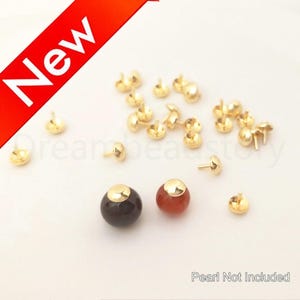 Op de afbeelding: Goudkleurige sieradenonderdelen, waaronder stud-palen en decoratieve doppen. Er worden ook twee edelsteen kralen getoond, een donkerbruine en een rode. Het woord "New" wordt weergegeven op een rode banner. "Pearl Not Included" staat onderaan.