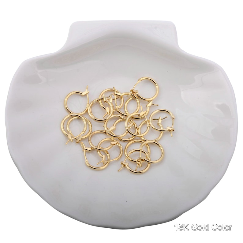 May include: A pile of gold-colored hoop earrings on a white seashell-shaped dish. 18K Gold Color.