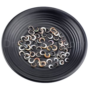Pode incluir: Uma coleção de pequenos amuletos de concha em forma de crescente em vários tons de marrom, branco e preto. Os amuletos estão dispostos em um prato preto.