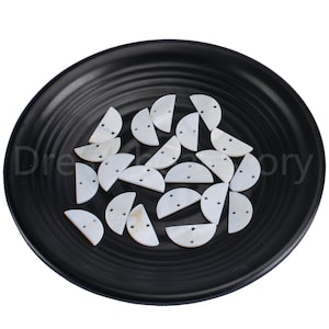 May include: A collection of white, half-circle shell buttons with two holes each, displayed on a black plate.