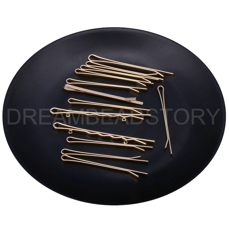 May include: A set of 15 gold hair clips on a black plate. The clips are various sizes and shapes, with some having a wavy design.