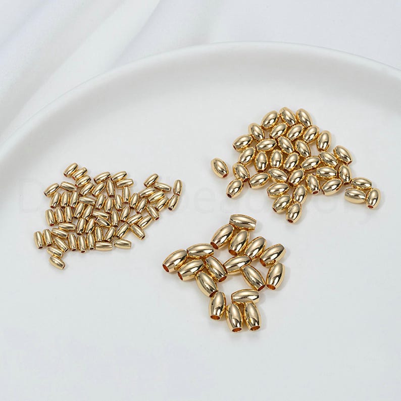 May include: A collection of small, gold-colored oval beads arranged in three separate groups. The beads have a smooth, reflective surface and are scattered on a white background. These beads are suitable for jewelry making and crafting.