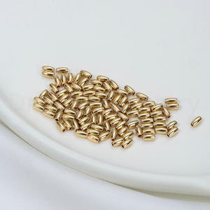 May include: A collection of small, gold-colored, oval-shaped beads. The beads are scattered on a white, slightly curved surface. The beads have a smooth, reflective surface, suggesting a metallic material.