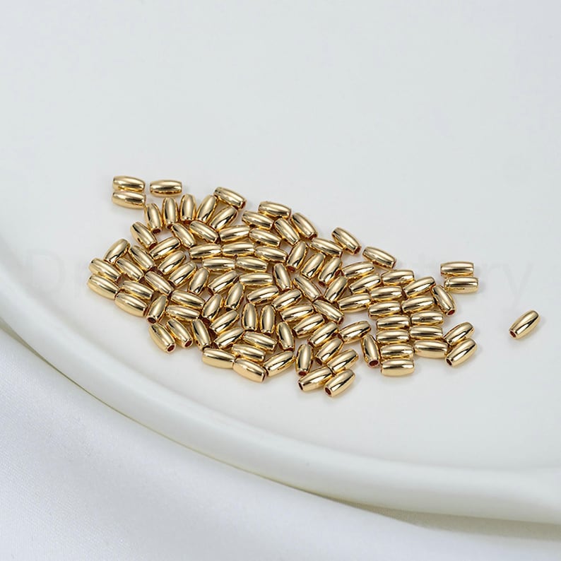 May include: A collection of small, gold-colored, oval-shaped beads. The beads are scattered on a white, slightly curved surface. The beads have a smooth, reflective surface, suggesting a metallic material.