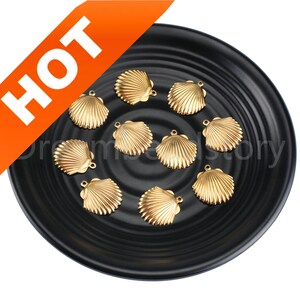 Può includere: Dieci ciondoli a forma di conchiglia in metallo dorato su un piatto nero. I ciondoli misurano circa 2,5 cm e hanno un design dettagliato.