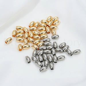 May include: A collection of oval-shaped beads in gold and silver tones. The beads are scattered on a white surface, with a soft, diffused light. These beads are suitable for jewelry making and crafting projects.