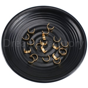 Puede incluir: Primer plano de un plato negro con 18 cierres en forma de U de metal dorado. Los cierres se utilizan para la fabricación de joyas.
