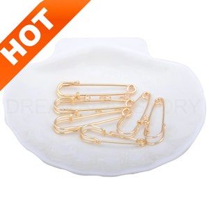 May include: A set of six gold safety pins in a white shell-shaped dish. The pins are arranged in a random pattern.