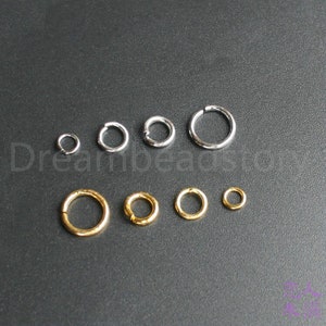 May include: Assortment of jump rings in silver and gold tones. The jump rings are different sizes and are arranged in a row on a gray surface.