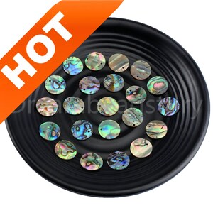 May include: A collection of 20 round iridescent abalone shell beads with two holes each. The beads are arranged on a black plate.