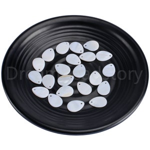 Puede incluir: Una colección de cuentas de concha blanca en forma de lágrima con un agujero en la parte superior. Las cuentas están dispuestas en un plato negro.