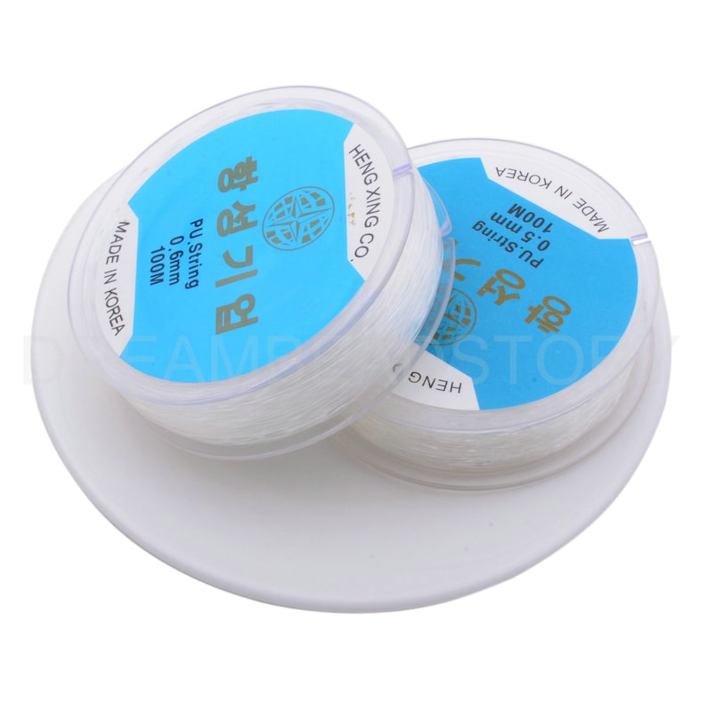 Puede incluir: Dos carretes de hilo el&aacute;stico transparente, cada uno con una etiqueta azul que dice "PU String 0.5 mm 100M" y "PU String 0.6 mm 100M" en coreano e ingl&eacute;s. Los carretes est&aacute;n sobre una base circular blanca.