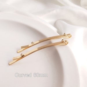 May include: Two gold-colored metal hair clips, each measuring 60 mm in length, are curved and have a smooth, shiny finish. The clips are arranged in a parallel position on a white surface. The text "Curved 60mm" is visible below the clips.