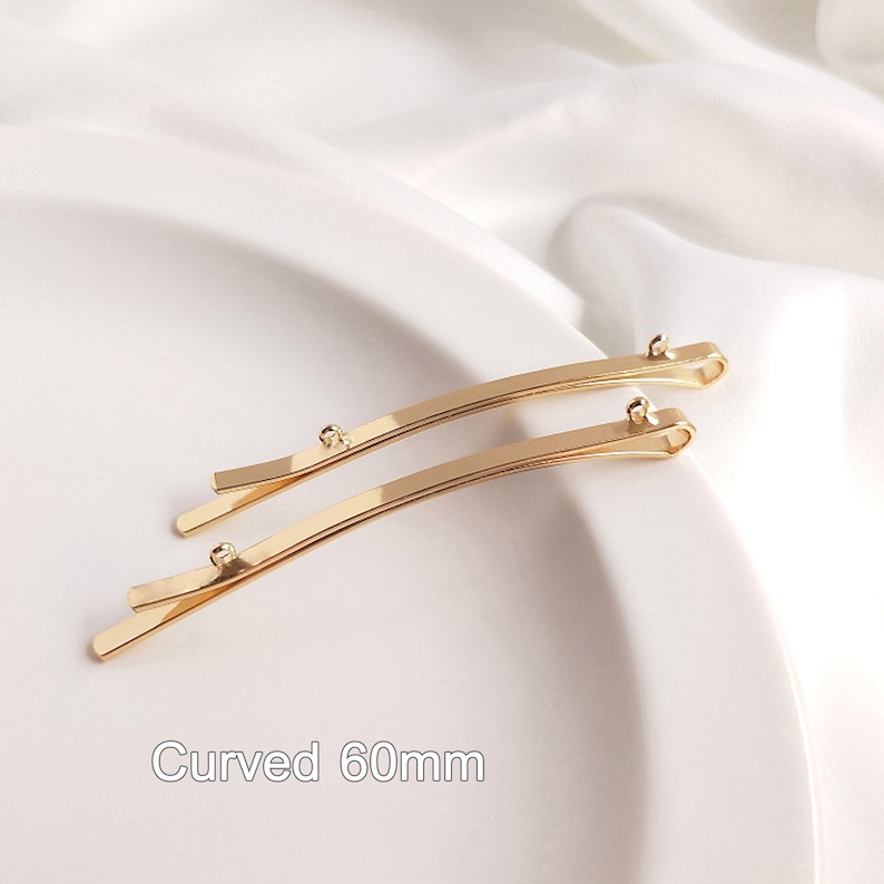 May include: Two gold-colored metal hair clips, each measuring 60 mm in length, are curved and have a smooth, shiny finish. The clips are arranged in a parallel position on a white surface. The text "Curved 60mm" is visible below the clips.