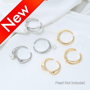 May include: A collection of open-ended rings in silver and gold tones, some with pearl accents. The rings have various designs, including a double-banded ring and a ring with a V-shaped design. The text "New" is visible in the upper left corner.