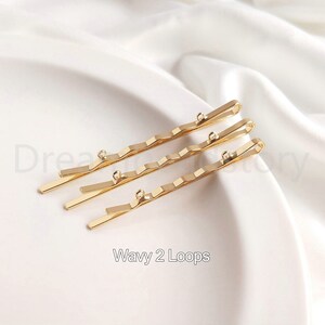May include: Three gold wavy hair clips with two loops at the end of each clip. The clips are arranged in a row, with the top clip slightly offset from the other two.