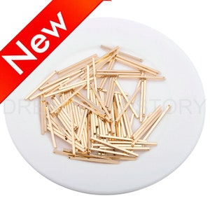 May include: A pile of gold-coloured metal bar connectors. The connectors are approximately 2.5 cm long and have a square cross-section. They are arranged in a random pattern on a white plate.