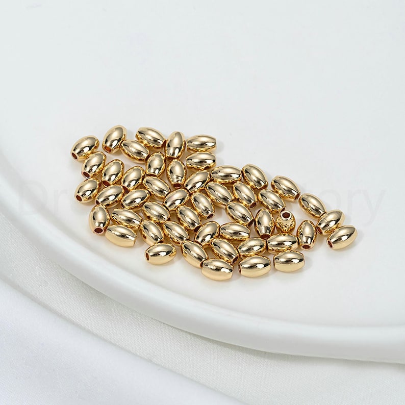 May include: A collection of small, gold-colored oval beads. The beads are scattered on a white, slightly curved surface. The beads have a smooth, reflective surface, suggesting a metallic material. The overall aesthetic is clean and minimalist.